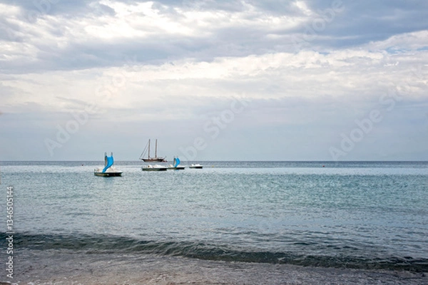 Obraz Beautiful picturesque view of the seashore and skyline at sunset. Sailboat and some pedalos moored in front of the beach with the horizon behind. 
