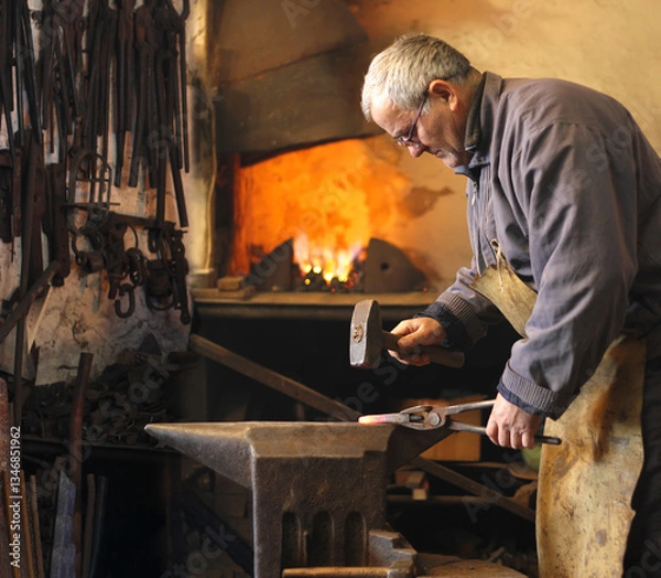 Obraz Blacksmith at work, mature man hitting a red-hot iron with a hammer on the anvil. Blacksmith forging red-hot metal bar in smithy