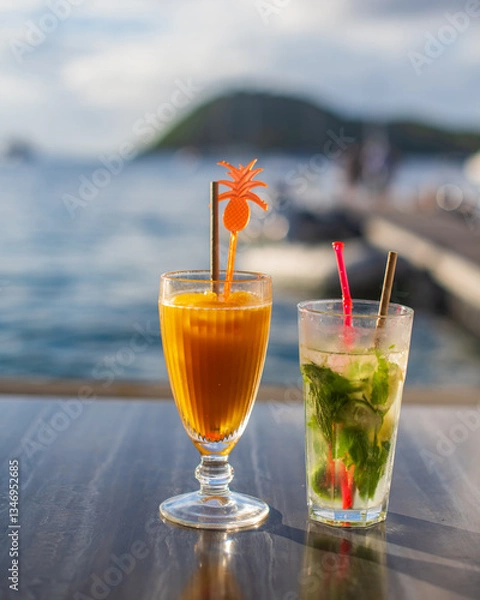 Fototapeta deux cocktails posés sur une table en bord de la baie des Saintes en Guadeloupe dans la mer des caraïbes avec un ponton en bois et une île à l'arrière plan.
