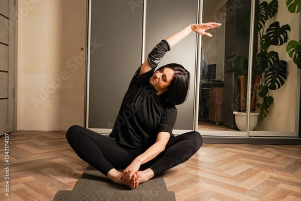 Fototapeta In the background is a wardrobe and a linoleum floor. A young woman in black sportswear is practicing yoga on a black mat in her room. The girl is in the lotus position.