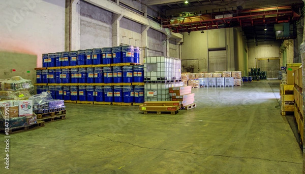 Fototapeta Blue barrels and other containers are neatly arranged on pallets in a spacious warehouse.Concrete floor and high ceiling give it an industrial feel,while the large open space allows for easy movement.