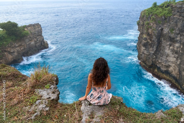 Fototapeta A beautiful woman in a pink dress sits on a cliff above the ocean on the island of Nusa Penida. Devil's Billabong an incredibly wonderful lagoon with splashes from the waves.