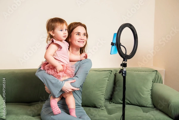 Obraz smiling mother holds her little daughter while recording a video with a smartphone and ring light.