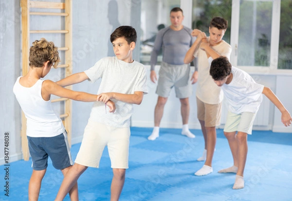 Fototapeta Focused teenage boys practicing close combat techniques in sparring in training room during self-defence workout under supervision of coach