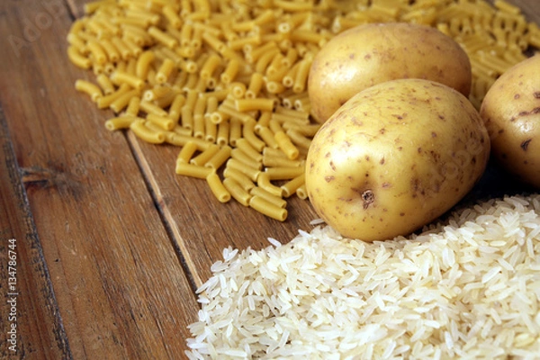 Obraz Rice, potatoes and macaroni pasta on a wooden table.Three common carbohydrates which provide energy but can cause obesity