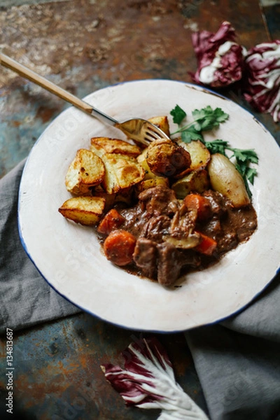 Fototapeta Beef bourguignon in ceramic plate with baked potatoes on metallic background. Rustic concept, natural light.