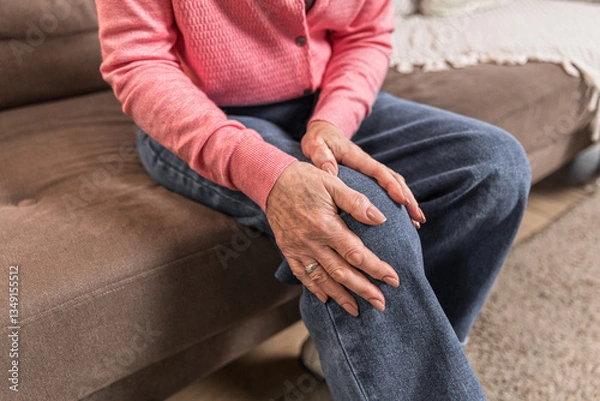Obraz Elderly woman feels sharp knee pain while standing up from couch at home. Her posture and frowning reflects discomfort, emphasizing the importance of joint health.