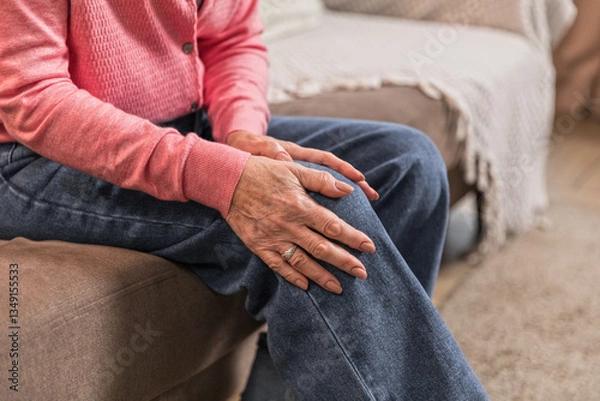 Obraz Elderly woman feels sharp knee pain while standing up from couch at home. Her posture and frowning reflects discomfort, emphasizing the importance of joint health.