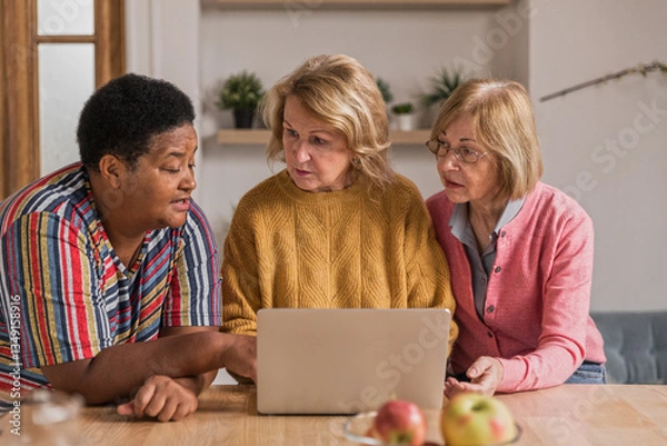 Obraz Senior Women Friends Gathering at Home. Three cheerful retired women enjoying friendly together in living room and chatting. Mature women use laptop computer typing on keyboard and pointing at screen