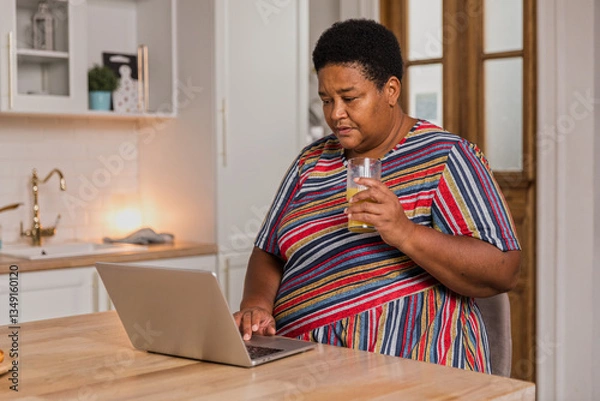 Obraz Senior afro woman sits at table typing on keyboard. Cheerful aged woman working remotely using laptop. Older focused housewife browsing online. Female retired enjoy using computer