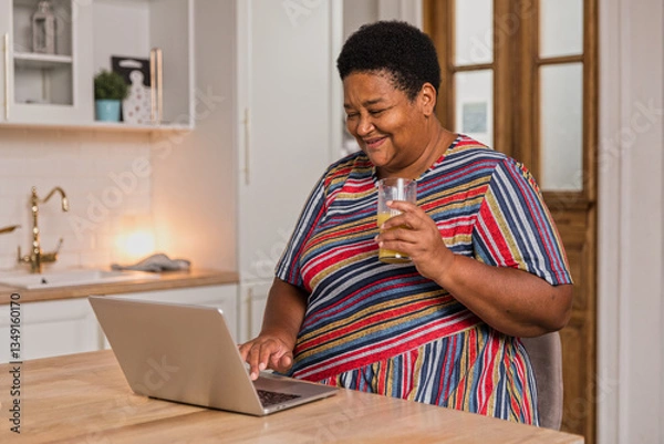 Obraz Senior afro woman sits at table typing on keyboard. Cheerful aged woman working remotely using laptop. Older focused housewife browsing online. Female retired enjoy using computer
