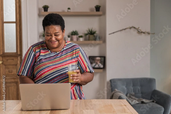 Obraz Senior afro woman sits at table typing on keyboard. Cheerful aged woman working remotely using laptop. Older focused housewife browsing online. Female retired enjoy using computer