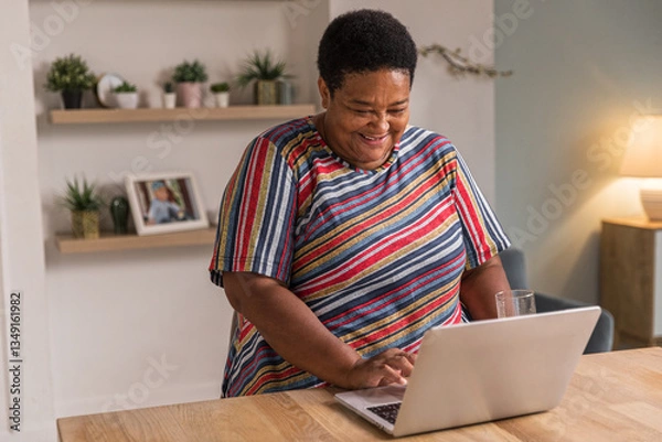 Obraz Senior afro woman sits at table typing on keyboard. Cheerful aged woman working remotely using laptop. Older focused housewife browsing online. Female retired enjoy using computer