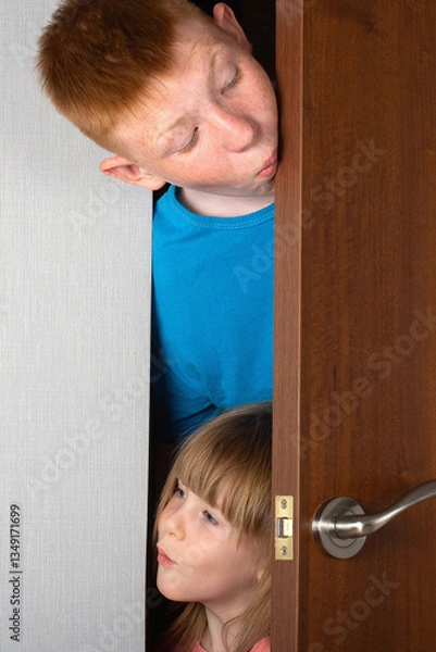 Obraz The children, a brother and a sister, peek curiously from behind the door of the room. vertical photo