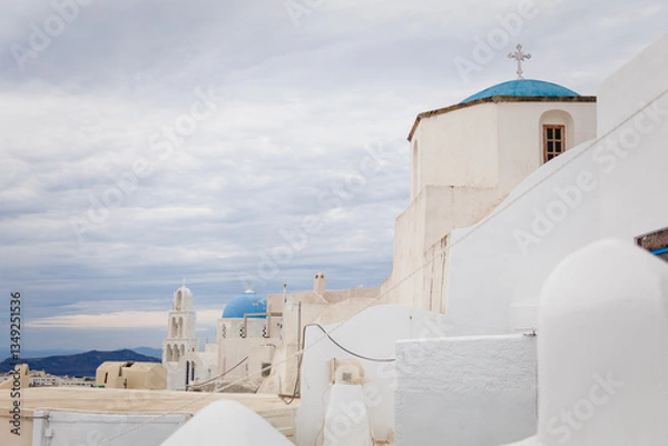 Fototapeta Horizontal view of white Greek orthodox church dome and tower and bells with cross in Pyrgos Kallistis, Santorini, Greece, with cloudy blue sky. A perfect day trip destination from Fira