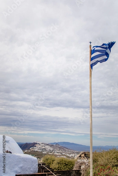 Fototapeta Vertical view from Pyrgos Kallistis, showcasing the Greek flag, cloudy sky, panoramic vistas of north Santorini, Aegean Sea, Greek islands, and coast, with whitewashed homes of Fira in the distance