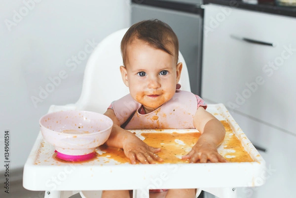 Fototapeta A smiling baby sits in a high chair covered in food, joyfully playing with puree while learning to eat independently in a modern kitchen