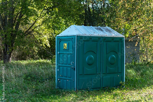 Fototapeta Green colored mobile public disabled toilet stands on the grass in the forest against a backdrop of trees.