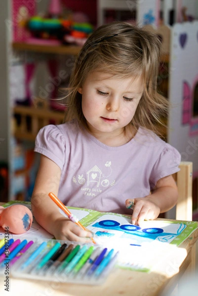 Fototapeta Kid paint. Child painting in sunny study room. Creative little artist at work. A charming girl is drawing with felt-tip pens at the table.