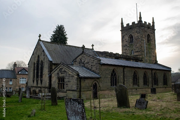 Fototapeta Eham, Chesterfield, United Kingdom - 21 Mar 2025 - Eham Parish church with a cemetery in front of it. The cemetery is full of headstones and the church is surrounded by trees. The sky is cloudy and th