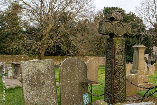 Fototapeta Eham, Chesterfield, United Kingdom - 21 Mar 2025 - A cemetery with a cross and several gravestones. The cross is in the middle of the cemetery and is surrounded by a chain link fence. The cemetery is 