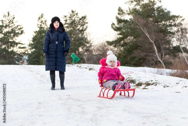 Obraz The child, a little girl riding on a sled with snow slides. Winter fun for children. A child with his mother and a winter walk.