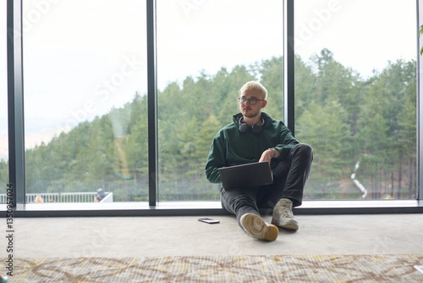Fototapeta A blonde man sitting on the floor of a modern office, leaning against the window with a laptop in his lap, fully engaged in his work while maintaining a relaxed and focused demeanor.