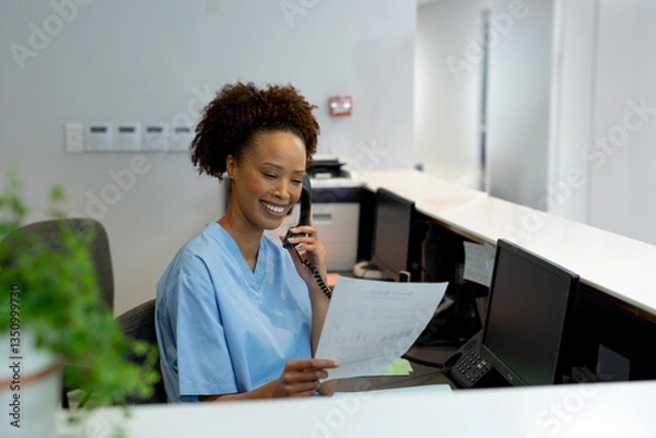 Fototapeta Hospital receptionist in scrubs smiling while talking on phone and holding document