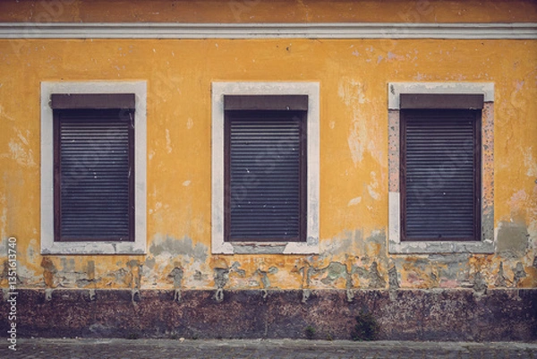 Fototapeta A weathered yellow wall with three closed shutters creates a striking geometric composition. The peeling paint and textured decay evoke a sense of time, history, and forgotten stories