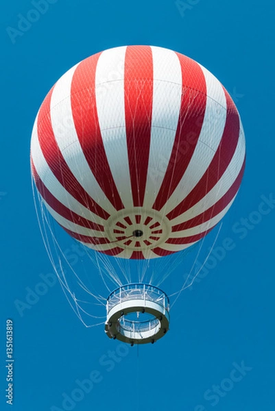 Fototapeta A striking red-and-white striped tethered balloon rising against a vibrant blue sky in Budapest’s City Park (Városliget). The perspective from below enhances the sense of height and adventure
