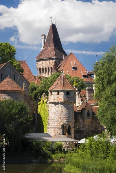 Fototapeta Vajdahunyad Castle in Budapest’s City Park blends medieval, Renaissance, and Baroque styles. Its towers and stone walls rise amid lush greenery, framed by ancient trees under a vivid blue sky