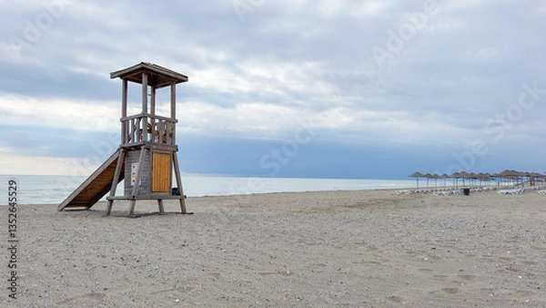Fototapeta Lifeguard tower on sandy empty beach with overcast cloudy sky and sea view. Wooden lifeguard tower. Serene coastal scene with ocean in the background. Safety concept