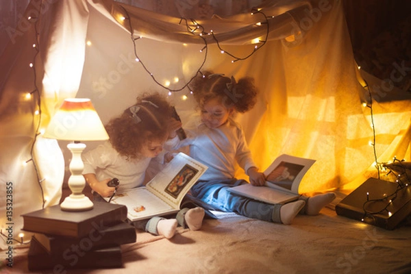 Fototapeta Reading and family games in children's tent. Two twins girls with books and flashlight before going to bed.