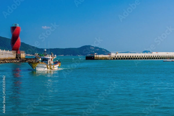 Fototapeta Fishing boat sailing near a vibrant red lighthouse in a harbor.