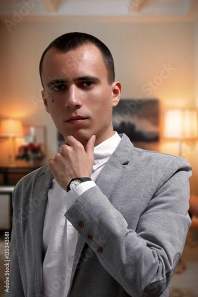 Obraz Portrait of goodlooking thoughtful young businessman in gray jacket and white t-shirt, looking at camera. Elegant interior on the background