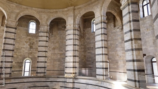 Fototapeta Pisa, Italy - December 20 2024. Interior of a stone rotunda of the Pisa Baptistry with striped marble columns, arched windows, and a circular walkway with metal railings.