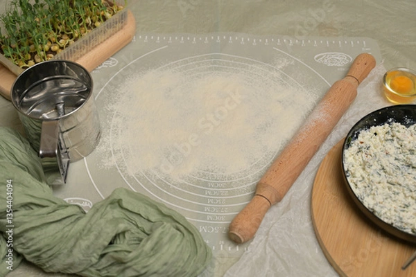 Obraz Floured silicone rug on worktop. Rolling pin, sieve, egg, green towel, microgreens sprouts, cottage cheese with parsley and dill greens on kitchen table. Home cooking baking