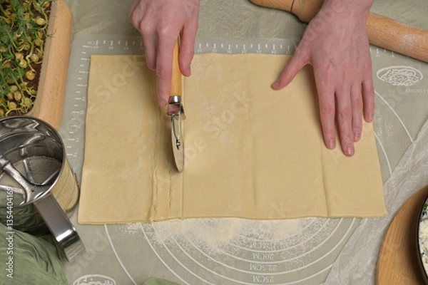 Obraz Woman's hands cuts a sheet of dough. Rolling pin, cutter, green towel, microgreens sprouts on kitchen table. Home cooking baking puffs