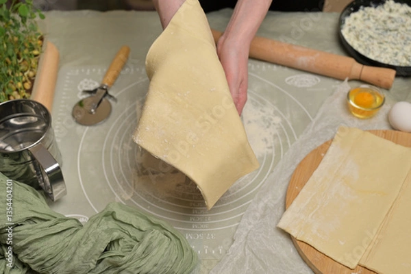 Obraz Woman's hands holding a sheet of dough. Rolling pin, cutter, eggs, green towel, microgreens sprouts on kitchen table. Home cooking baking puffs