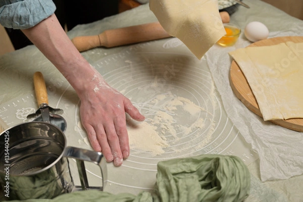 Obraz Woman's hand holding a sheet of dough and sprinkles flour worktop. Rolling pin, cutter, eggs, green towel on kitchen table. Home cooking baking puffs