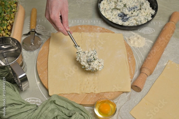 Obraz Woman's hand with fork putting cottage cheese with parsley and dill greens on dough. Rolling pin, cutter, egg, green towel and microgreens sprouts on kitchen table. Home cooking baking puffs