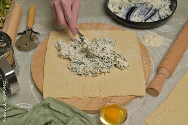 Obraz Woman's hand with fork putting cottage cheese with parsley and dill greens on dough. Rolling pin, cutter, egg, green towel and microgreens sprouts on kitchen table. Home cooking baking puffs