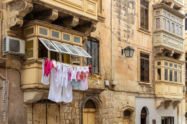 Obraz Traditional Maltese balcony with drying laundry. Historic limestone buildings with wooden enclosed balconies in Malta, featuring colorful clothes hanging out to dry, showcasing local charm.
