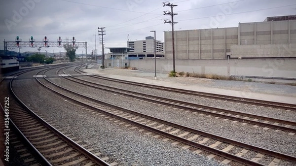 Fototapeta Multiple curving railway tracks with signal lights, along with concrete barriers and a parking structure visible in the background under an overcast sky