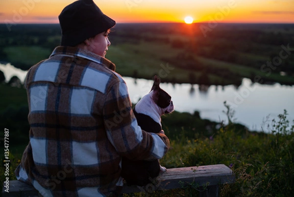 Fototapeta A boy sits on a bench and communicates with his friend, a Boston Terrier dog, in nature at sunset in summer.