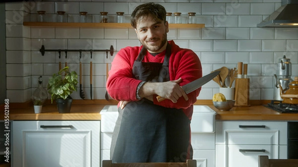 Obraz Confident man in modern kitchen holding sharp knife, wearing black apron and red sweater, smiling with professional chef-like attitude