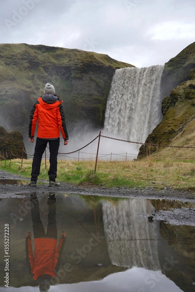 Fototapeta A hiker stands in a landscape with a stunning waterfall in Iceland. Rear view. Dressed in a hiking outfit and hat. Reflected in a puddle.