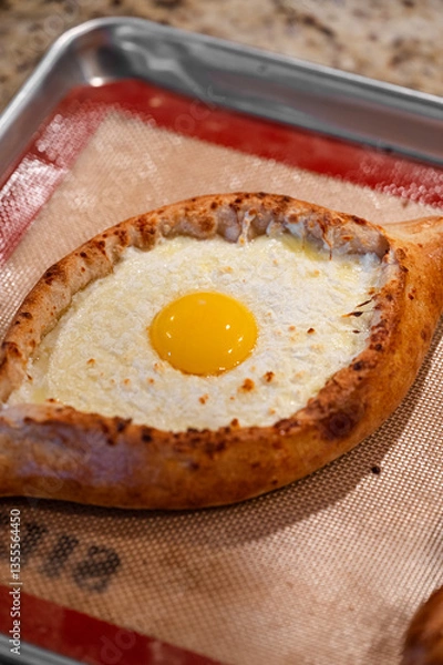 Obraz Traditional Adjarian Khachapuri filled with melted cheese and topped with a sunny egg yolk, served hot on a baking tray with a silicone mat