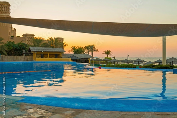 Fototapeta Luxury hotel pool in Sharjah at sunset, with golden light reflecting on the water. Elegant sun loungers line the poolside, offering a serene view of the beach and tranquil sea waves.
