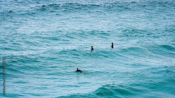 Fototapeta Surfers ride the waves in a vibrant ocean under a cloudy sky at midday